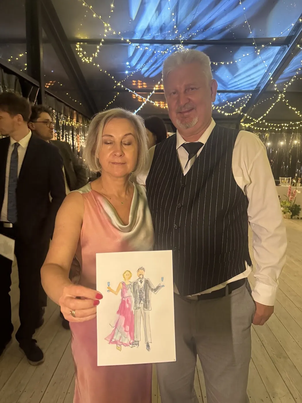 Starsza para gości pozuje z akwarelową ilustracją pod girlandą świateł