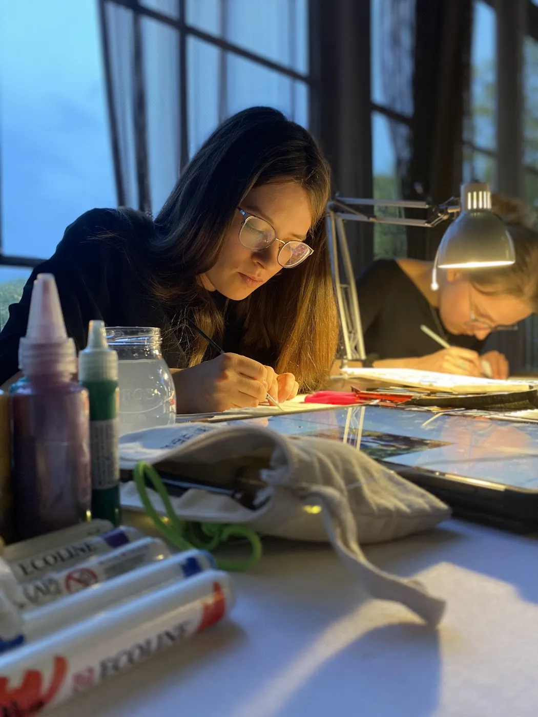Artystka skupiona nad akwarelowym portretem przy lampce, obok druga ilustratorka
