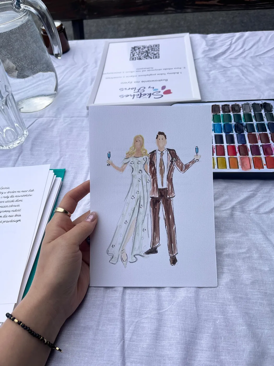Akwarelowa ilustracja pary gości trzymana obok palety farb w Dębowym Dworze